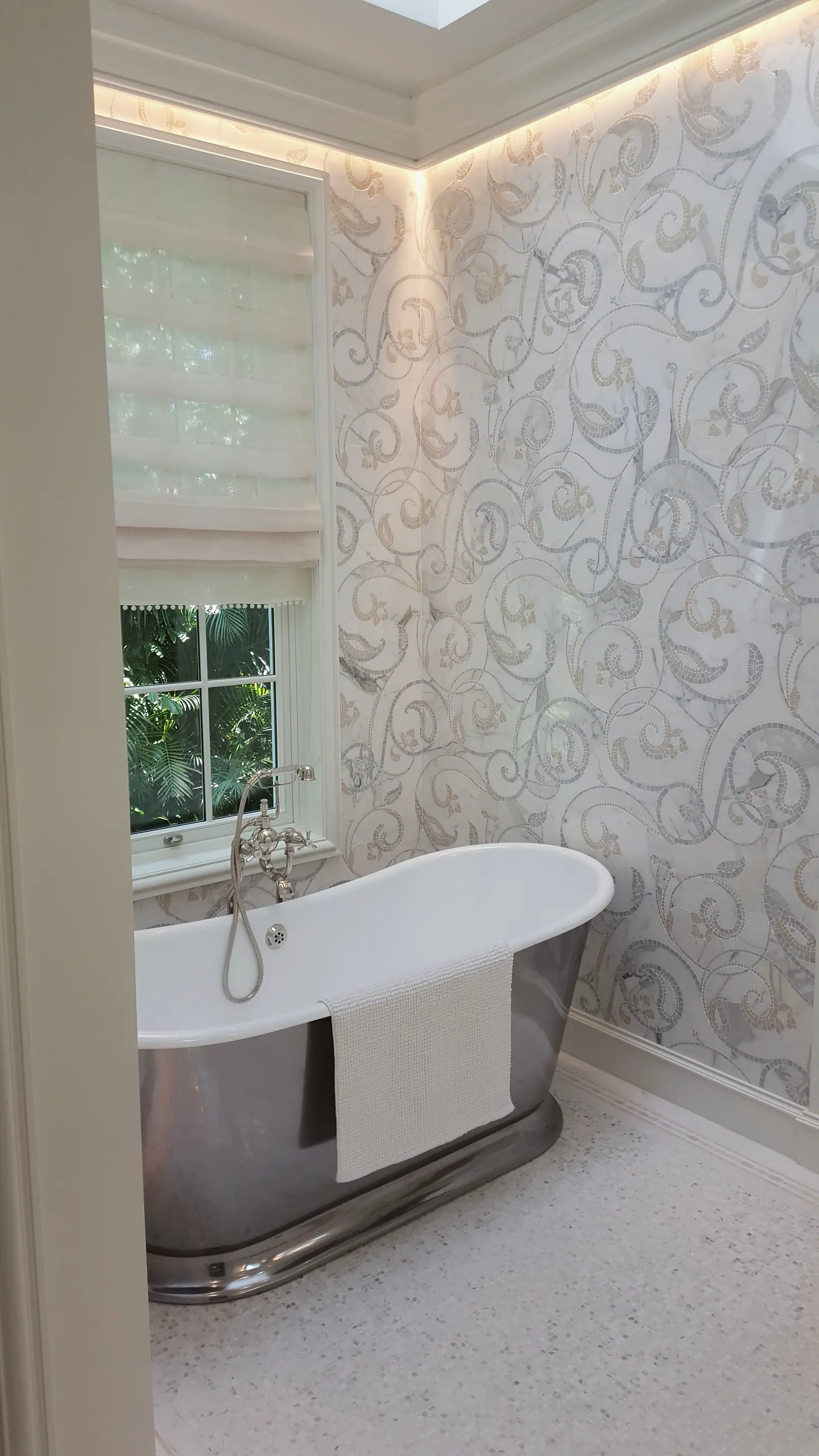 Freestanding tub with polished metal skirt in front of a scrolling marble mosaic wall, cove lit ceiling and a shaded window