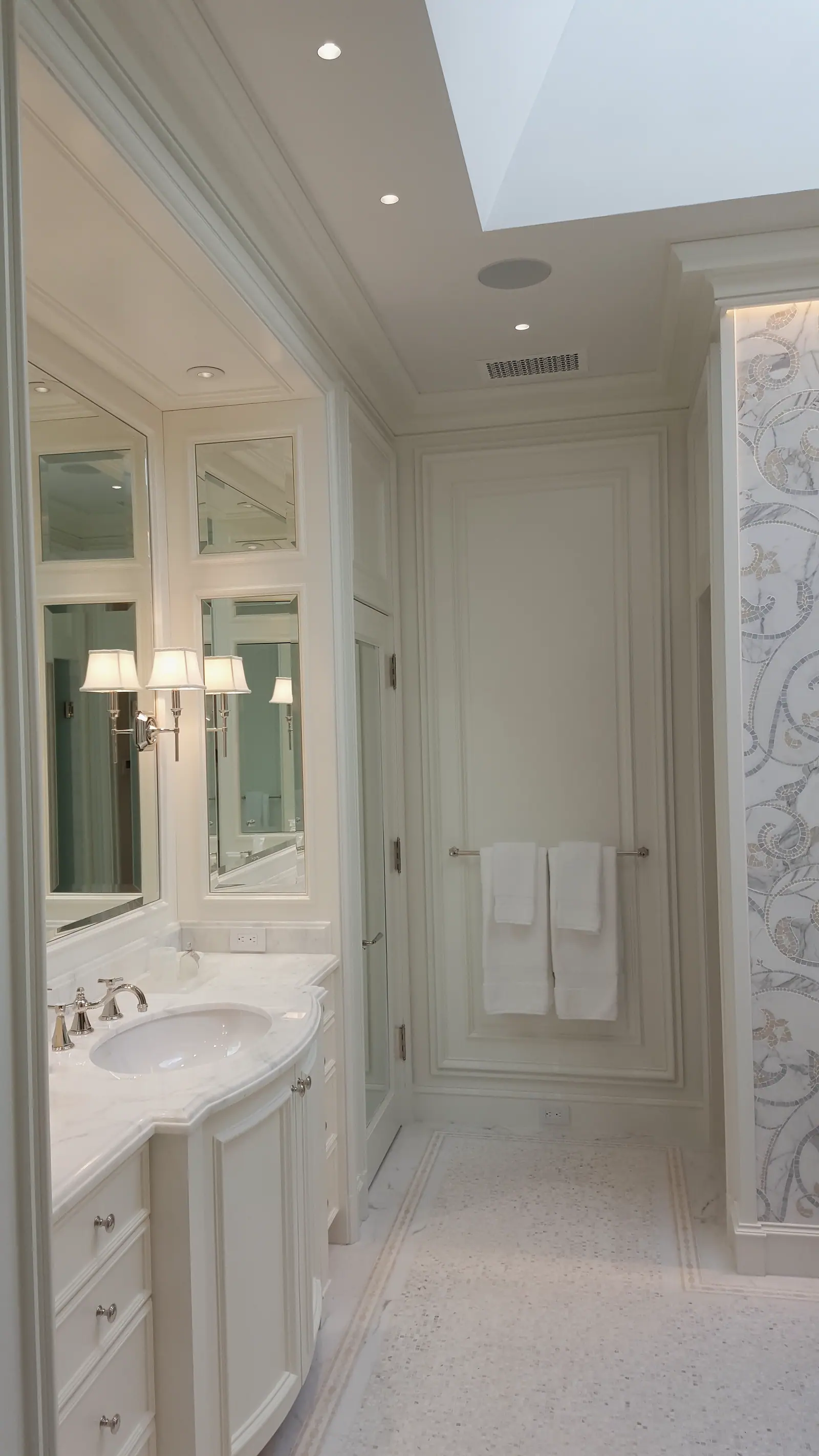Curved white vanity with marble top, mirrored millwork and sconces, paneled walls, skylight and marble mosaic floor