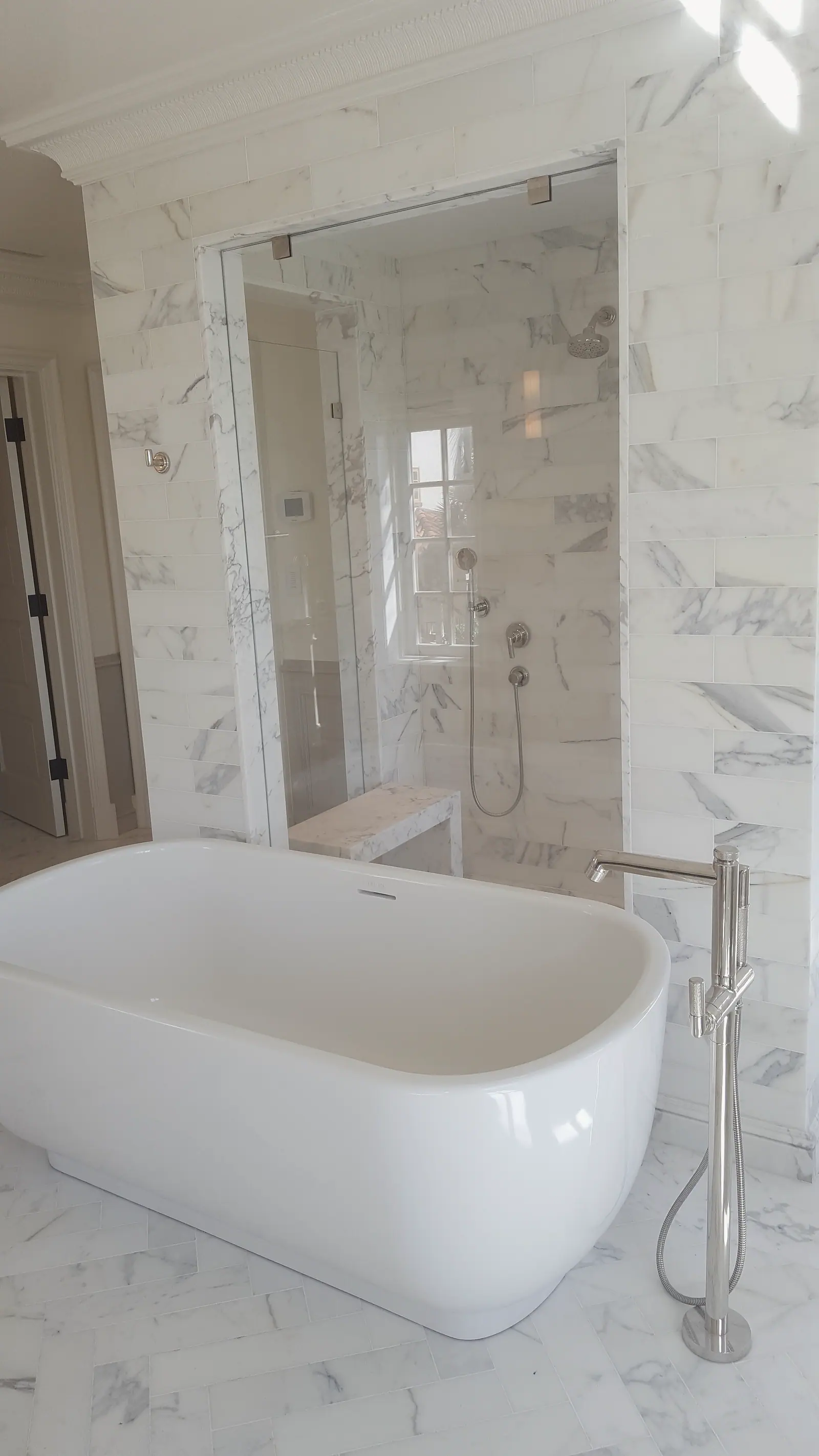Freestanding soaking tub and floor mounted filler before a marble clad glass shower with bench on marble herringbone floor
