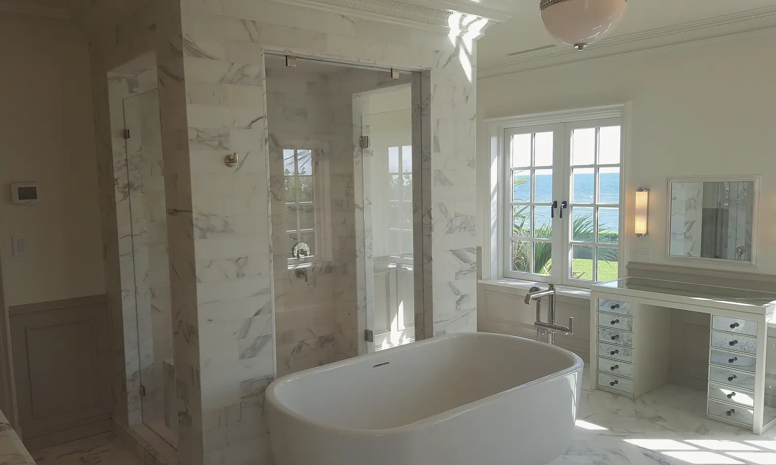 Primary bath with freestanding soaking tub, white marble shower with frameless glass, mirrored vanity and French windows with an ocean view