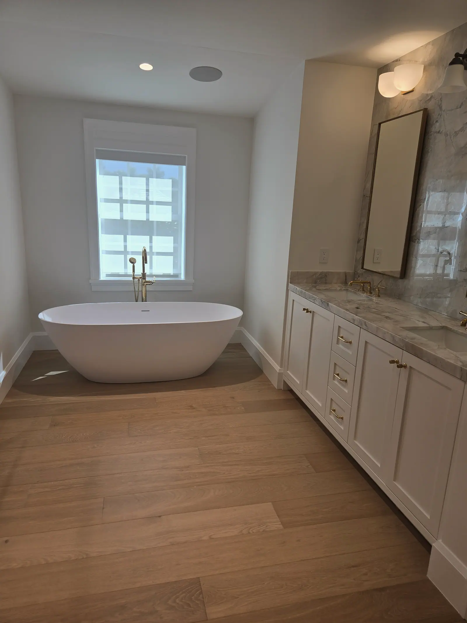 Freestanding tub on white oak floor beside a white double vanity with marble top and brass faucets