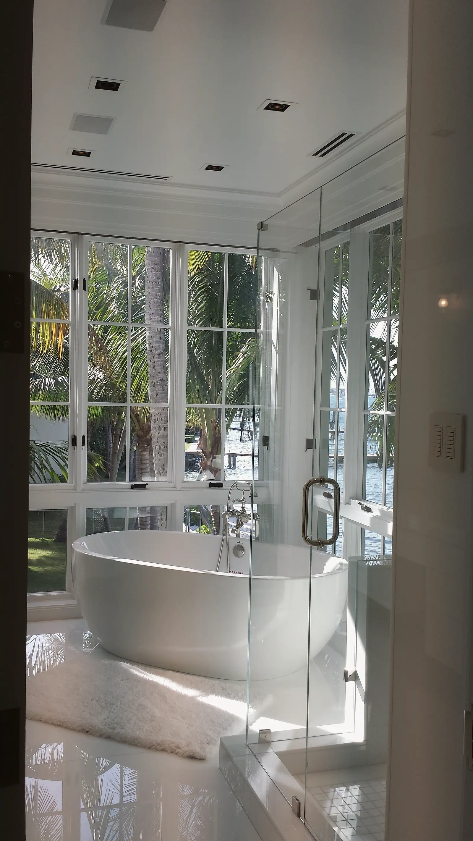 Freestanding soaking tub in a corner of tall windows overlooking palms and water, beside a frameless glass shower