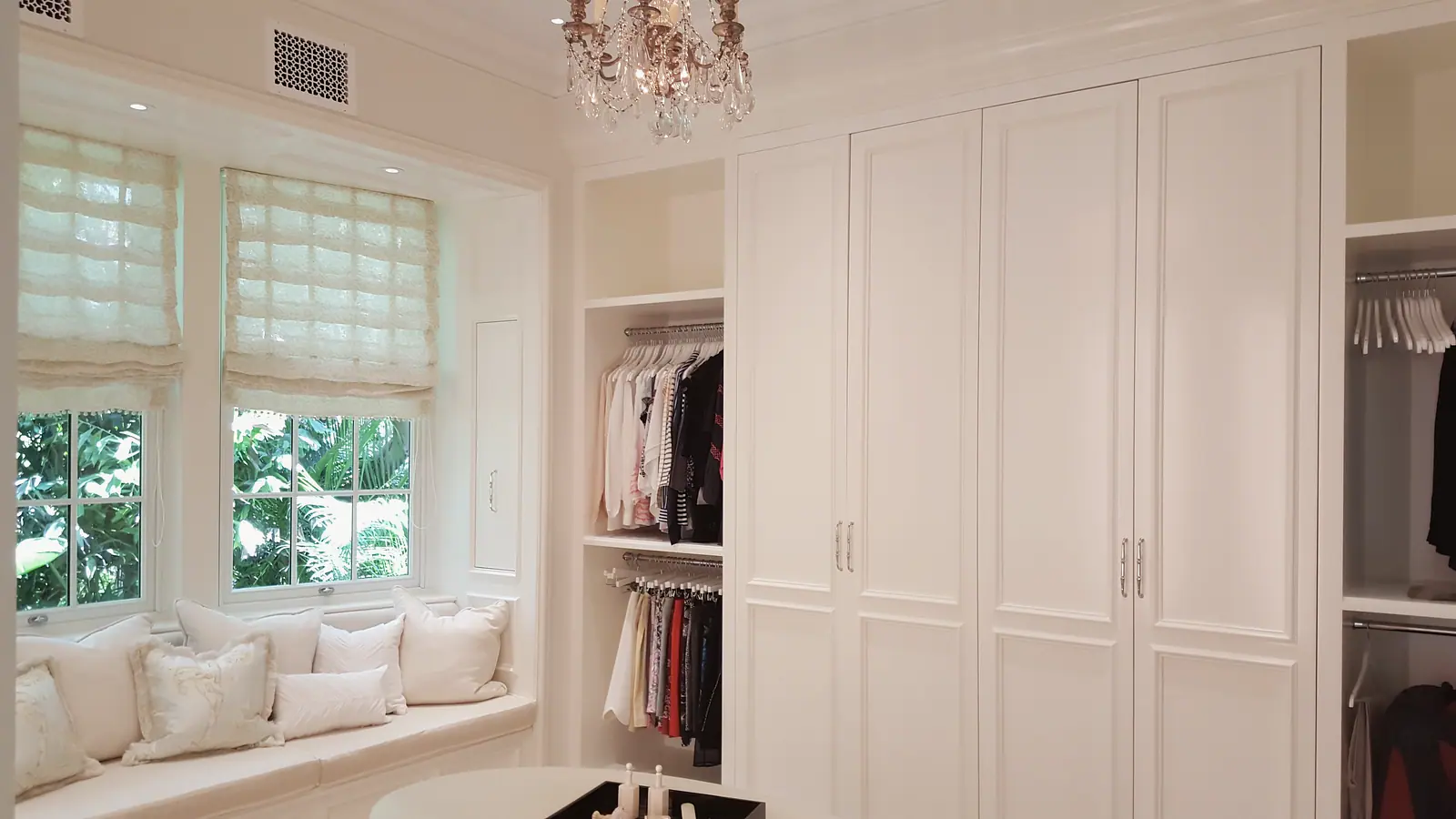 Dressing room with cushioned window seat under Roman shades, crystal chandelier and white paneled wardrobe cabinetry