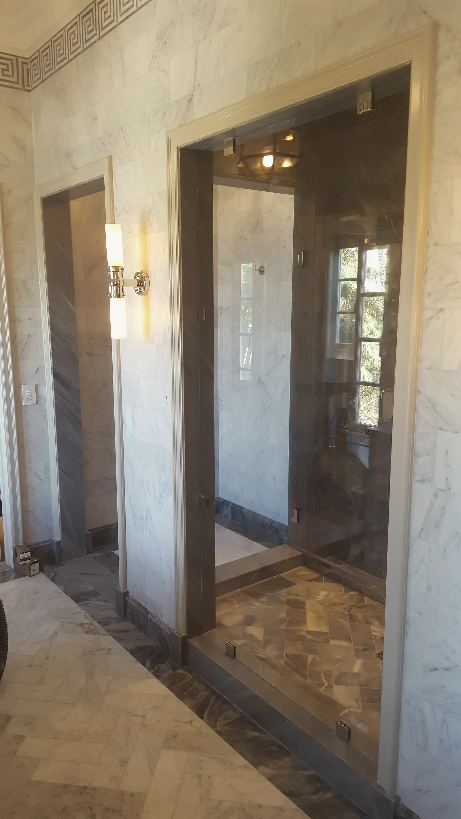 Frameless glass shower in white marble wall with gray marble floor, Greek key border and nickel sconce