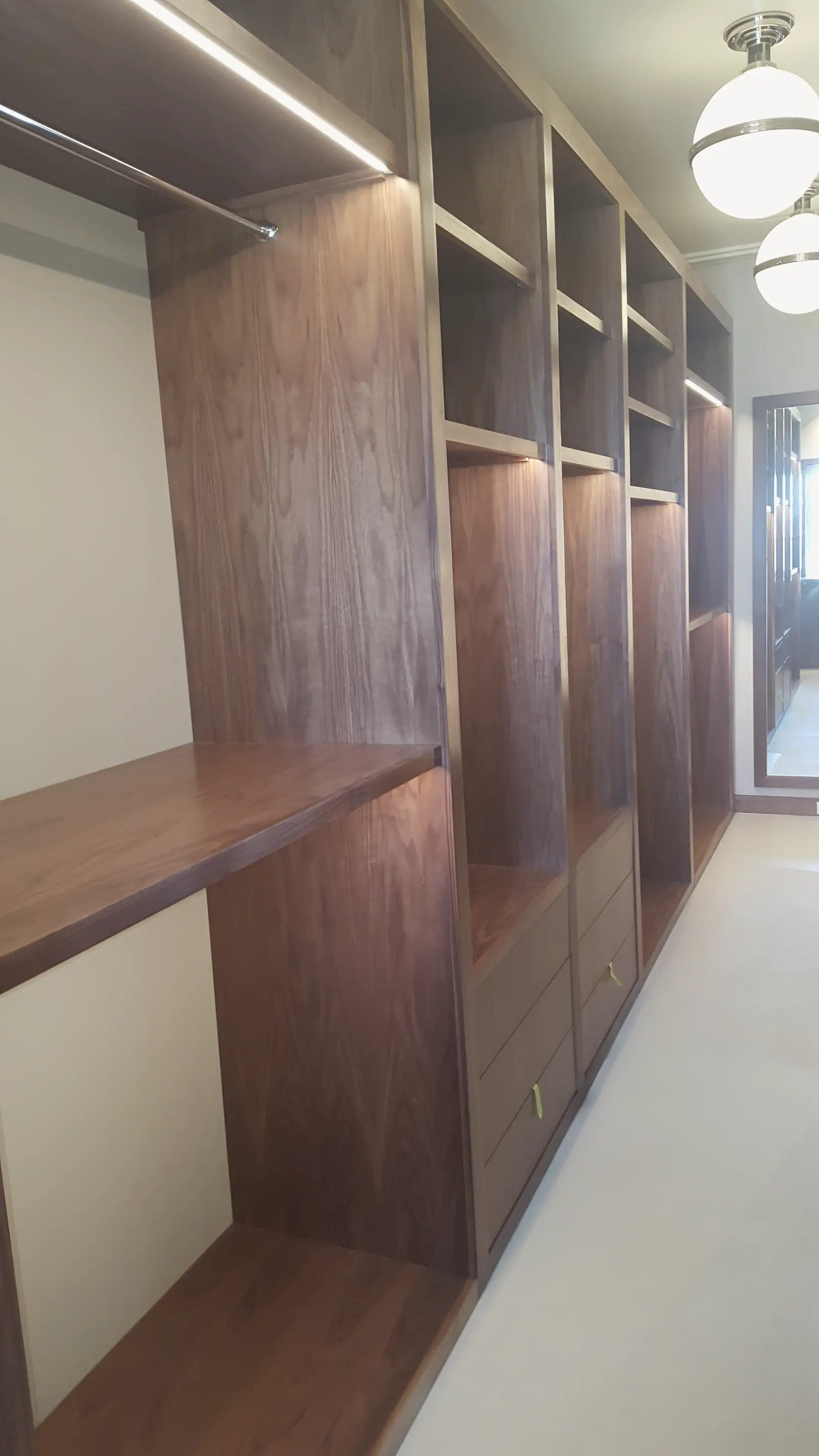 Walnut walk-in closet with lit shelving, hanging rod, drawers and globe pendant lights