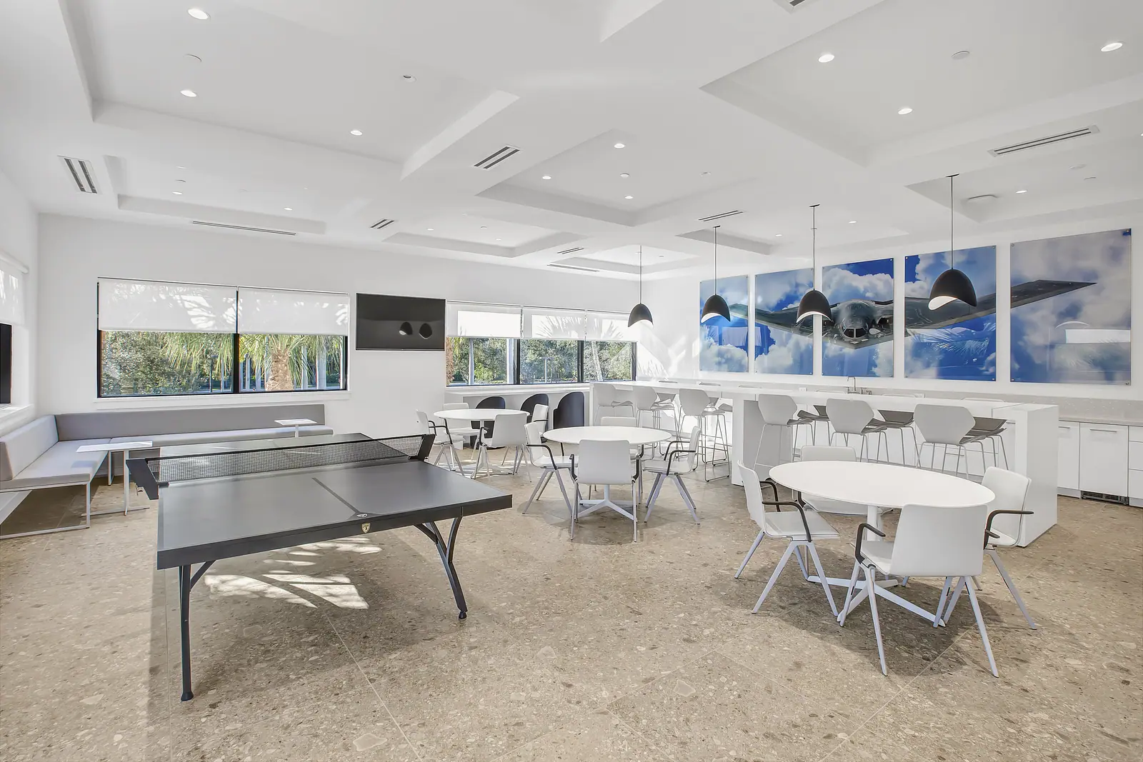 Office break room with coffered ceiling, ping pong table, round cafe tables and a long counter with stools under black pendants