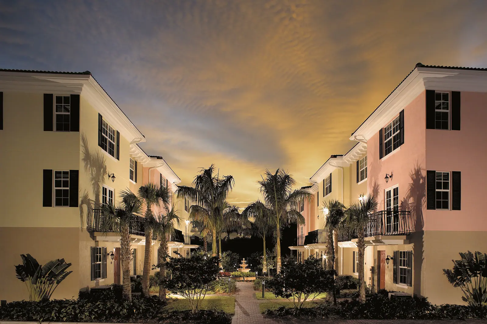 Pastel three story townhomes around a palm lined courtyard with a fountain, lit at dusk under a glowing sky