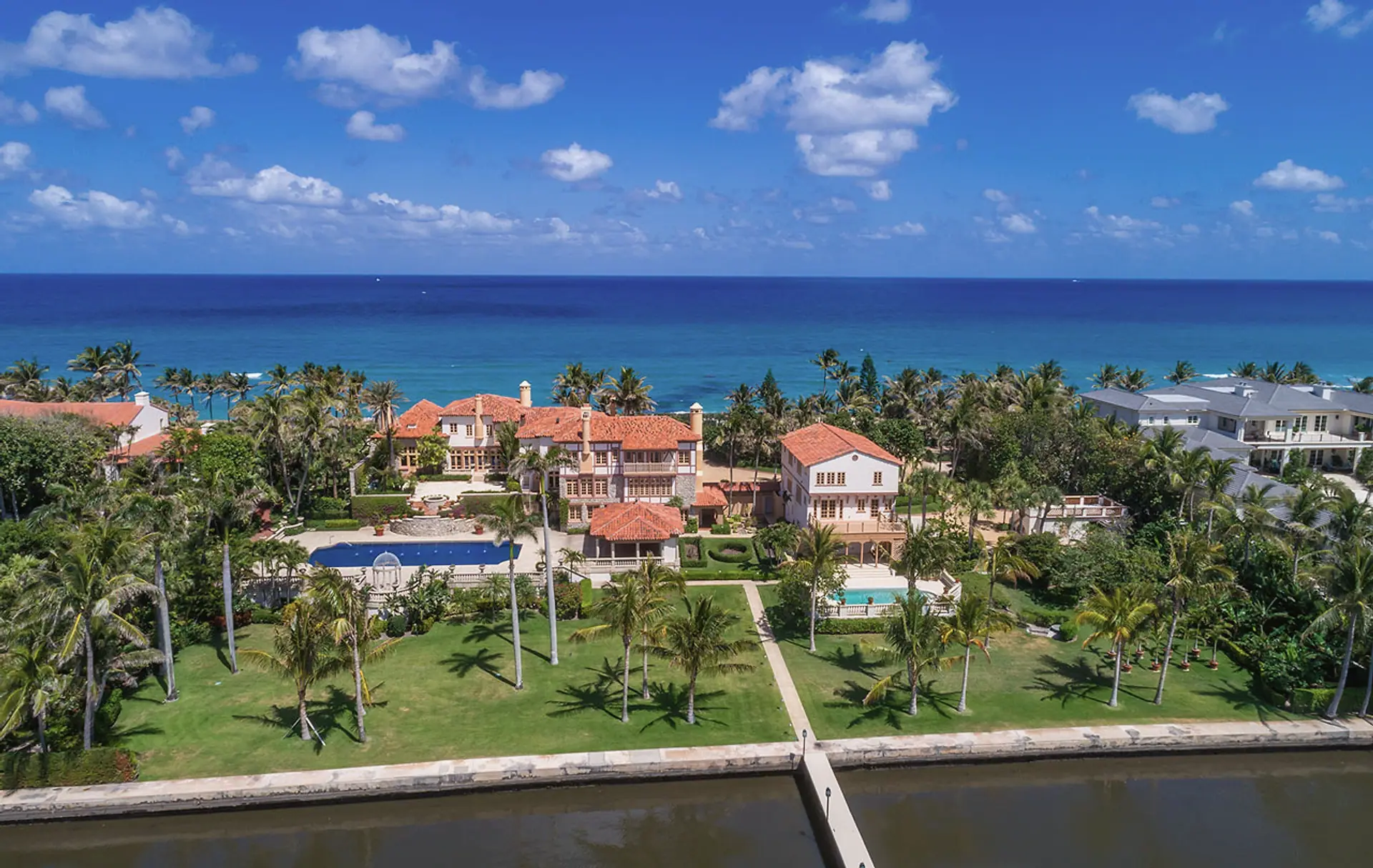 Aerial view of an oceanfront estate with red tile roofs and a long lawn running down to the water