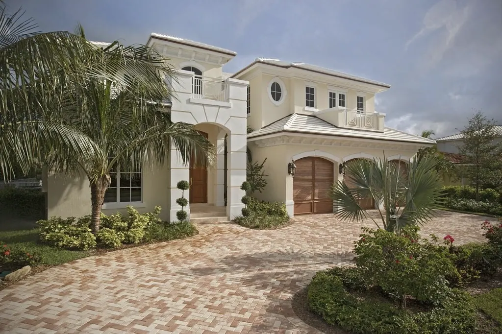 Pale stucco house with an arched entry tower, wood garage doors and a brick paver drive