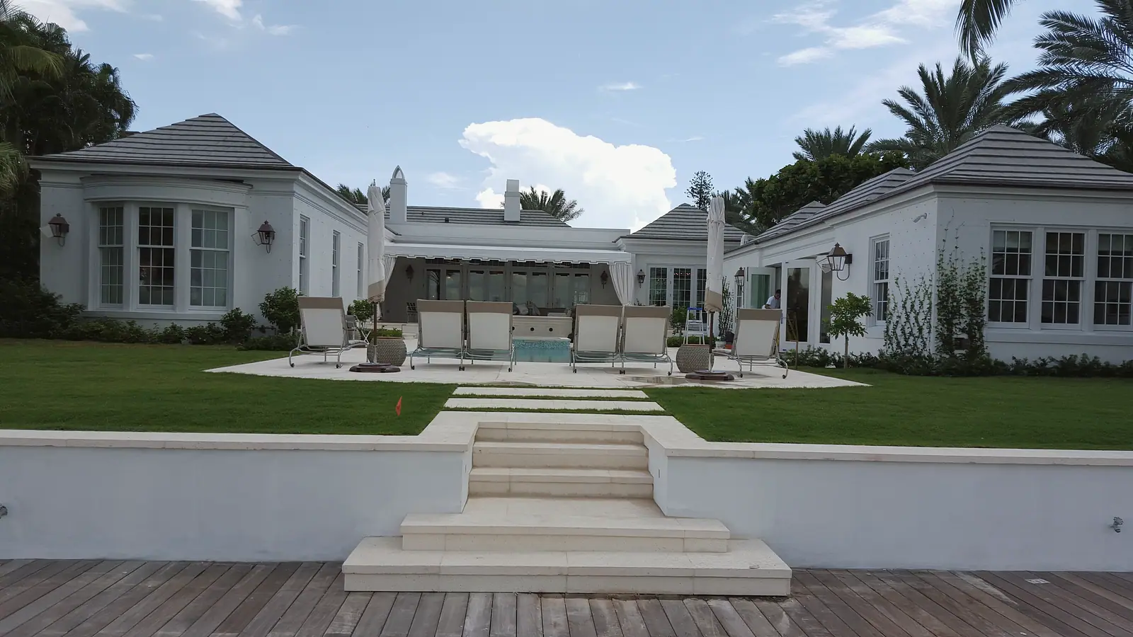 Rear of a white estate with gray tile hipped roofs, pool terrace with lounge chairs and lawn, stone steps rising from a wood deck