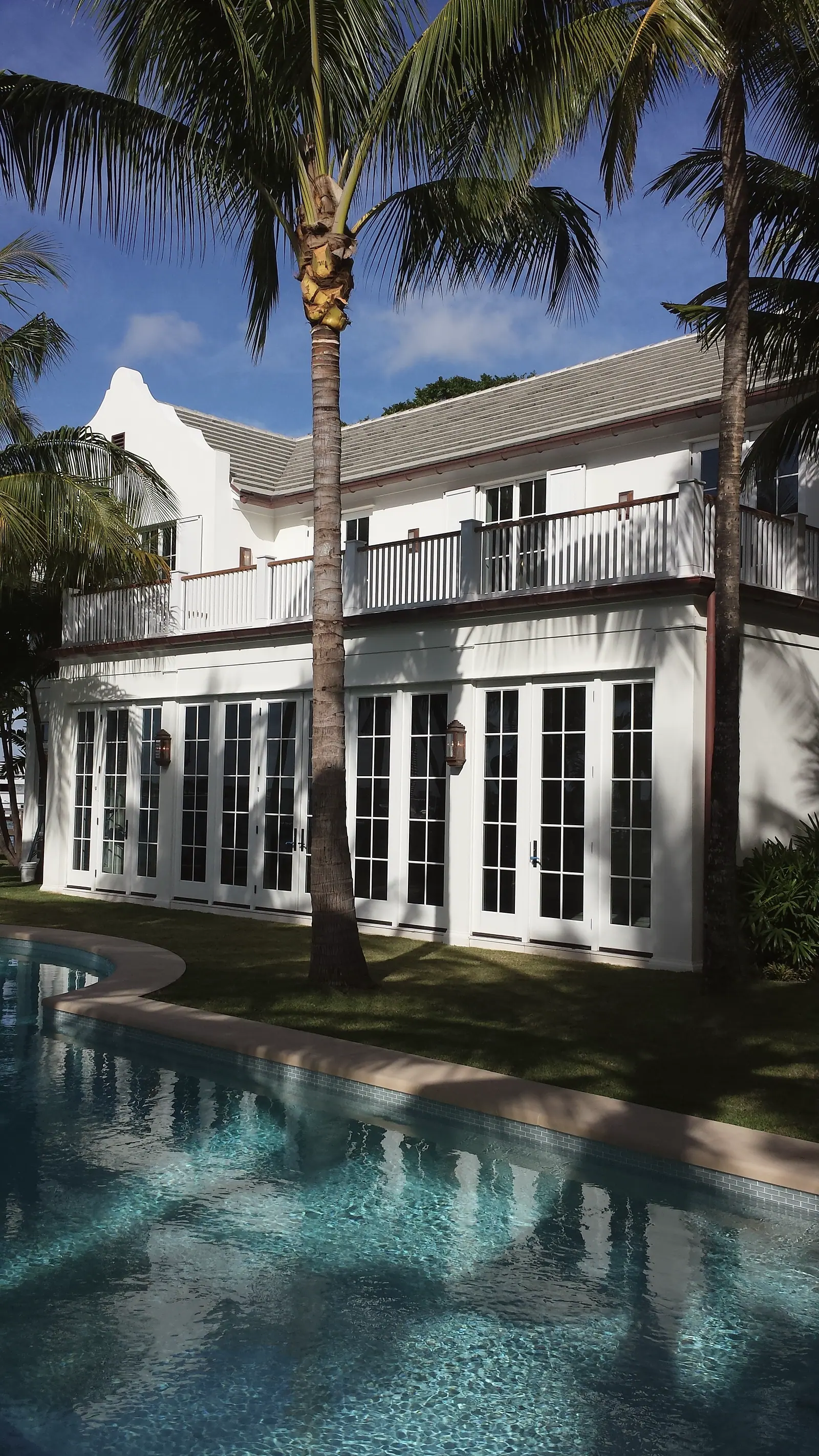 White two-story house with Dutch gable, upper balcony and a row of French doors behind a curved pool and palms