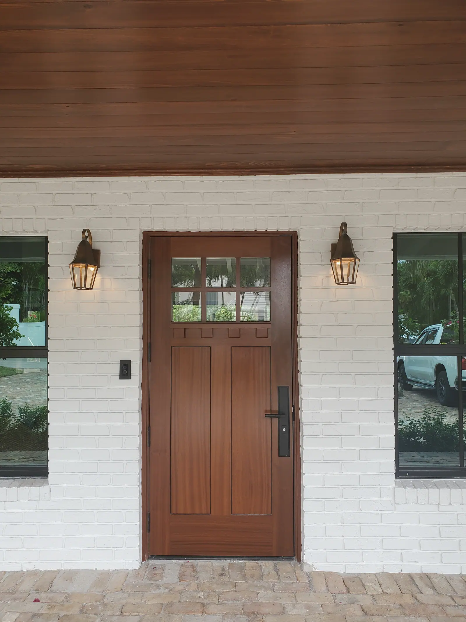 Mahogany front door between copper lanterns on white painted brick under a stained tongue and groove porch ceiling