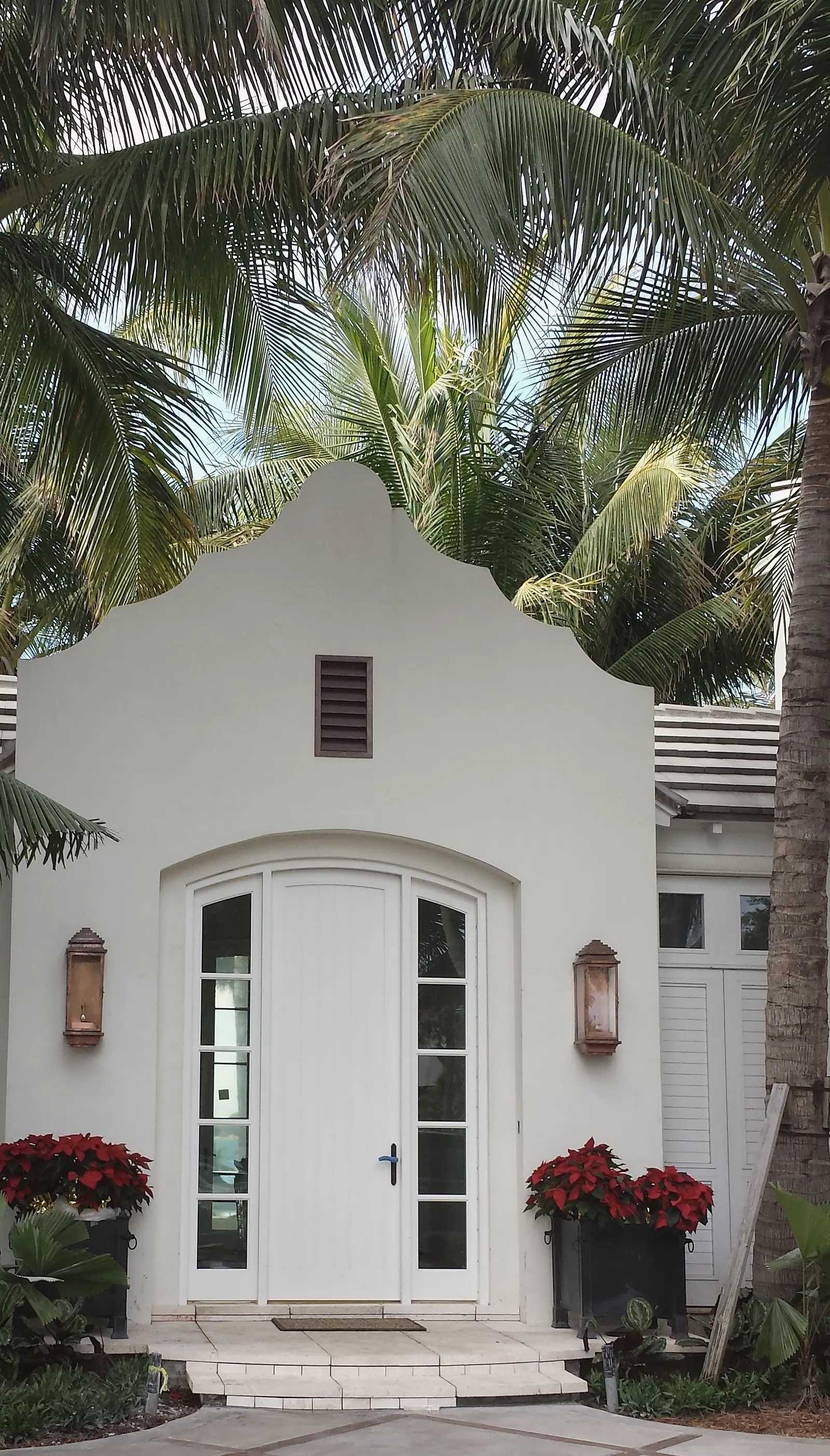 White Dutch gable entry with arched door and sidelights, copper lanterns and planters under coconut palms