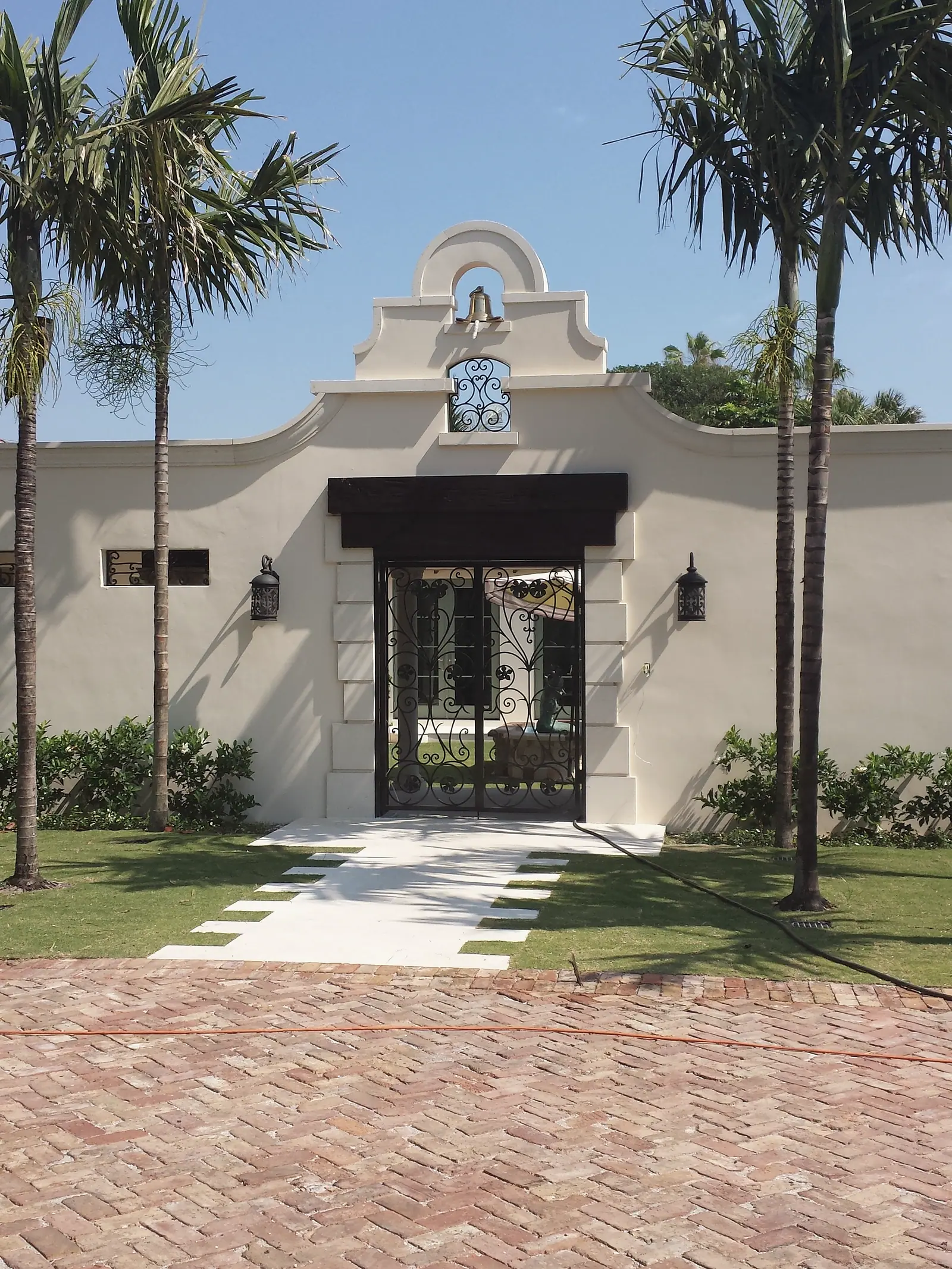 Centered bell gable entry wall with wrought iron gate, lanterns and a stone path across the lawn to a brick drive