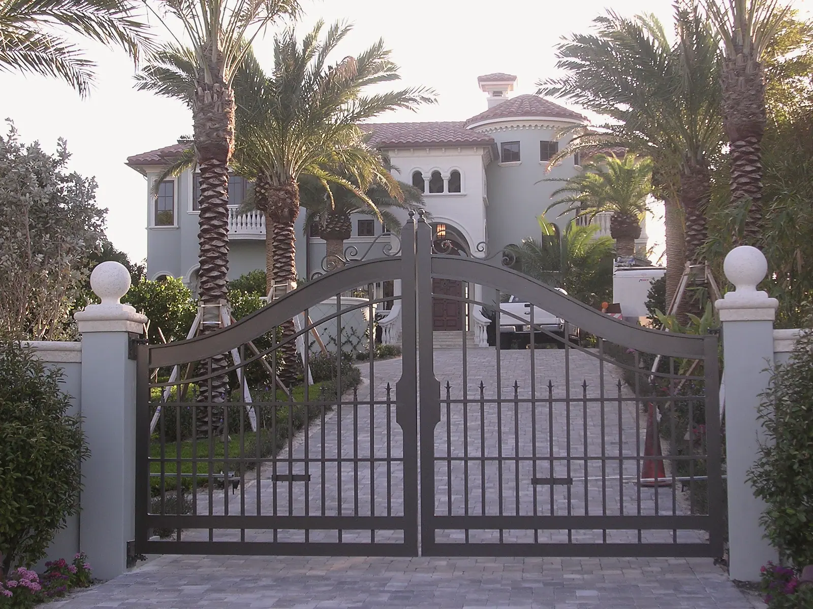 Mediterranean style house with a clay tile roof and round tower behind arched iron driveway gates and a paver drive