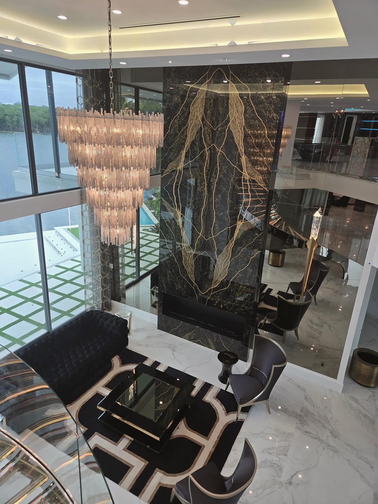 Double height living room seen from above with a black and gold stone fireplace wall, tiered crystal chandelier and marble floors