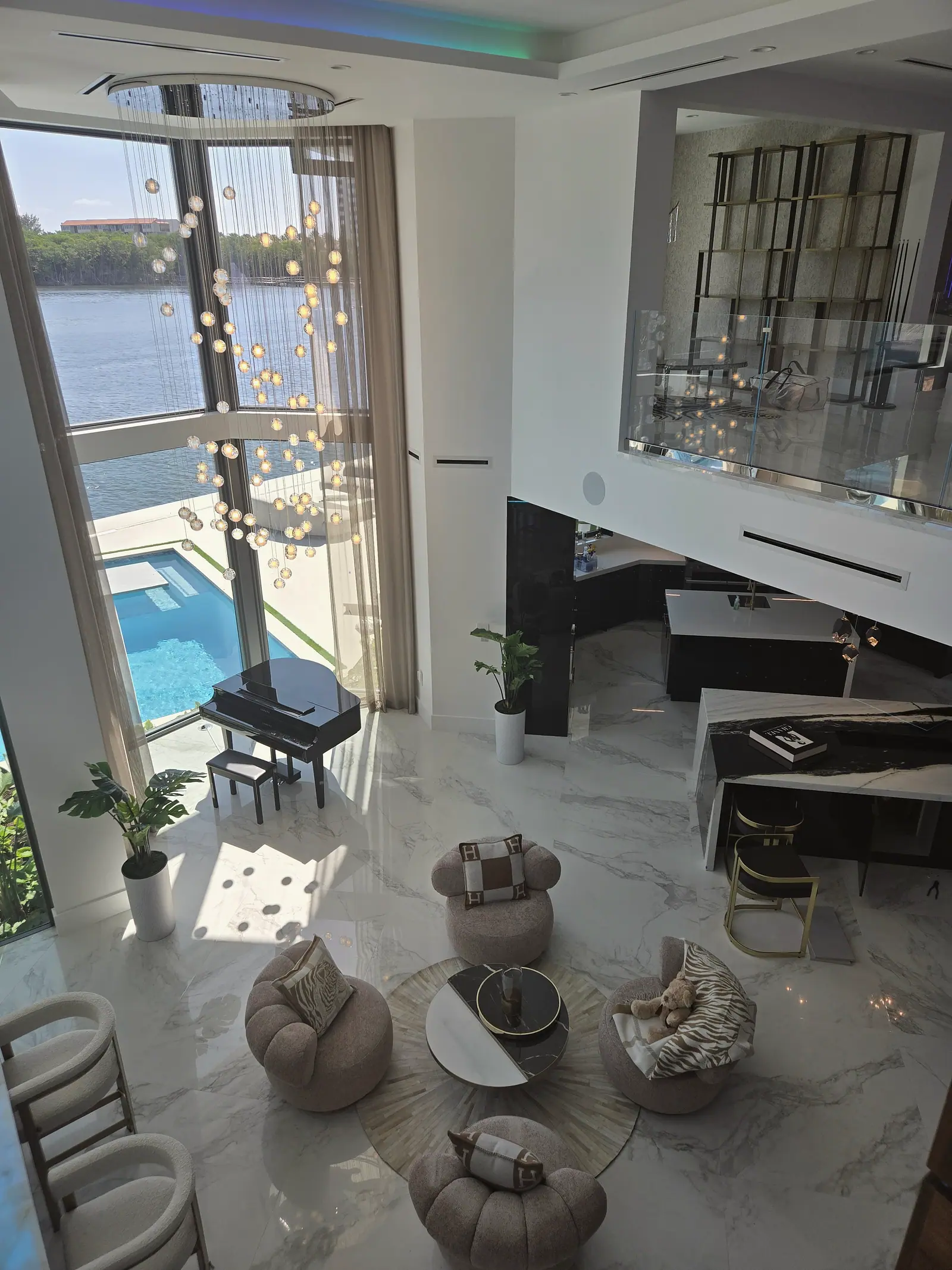 Double height living room from above with a cascading globe chandelier, piano by tall windows, pool outside and an open kitchen
