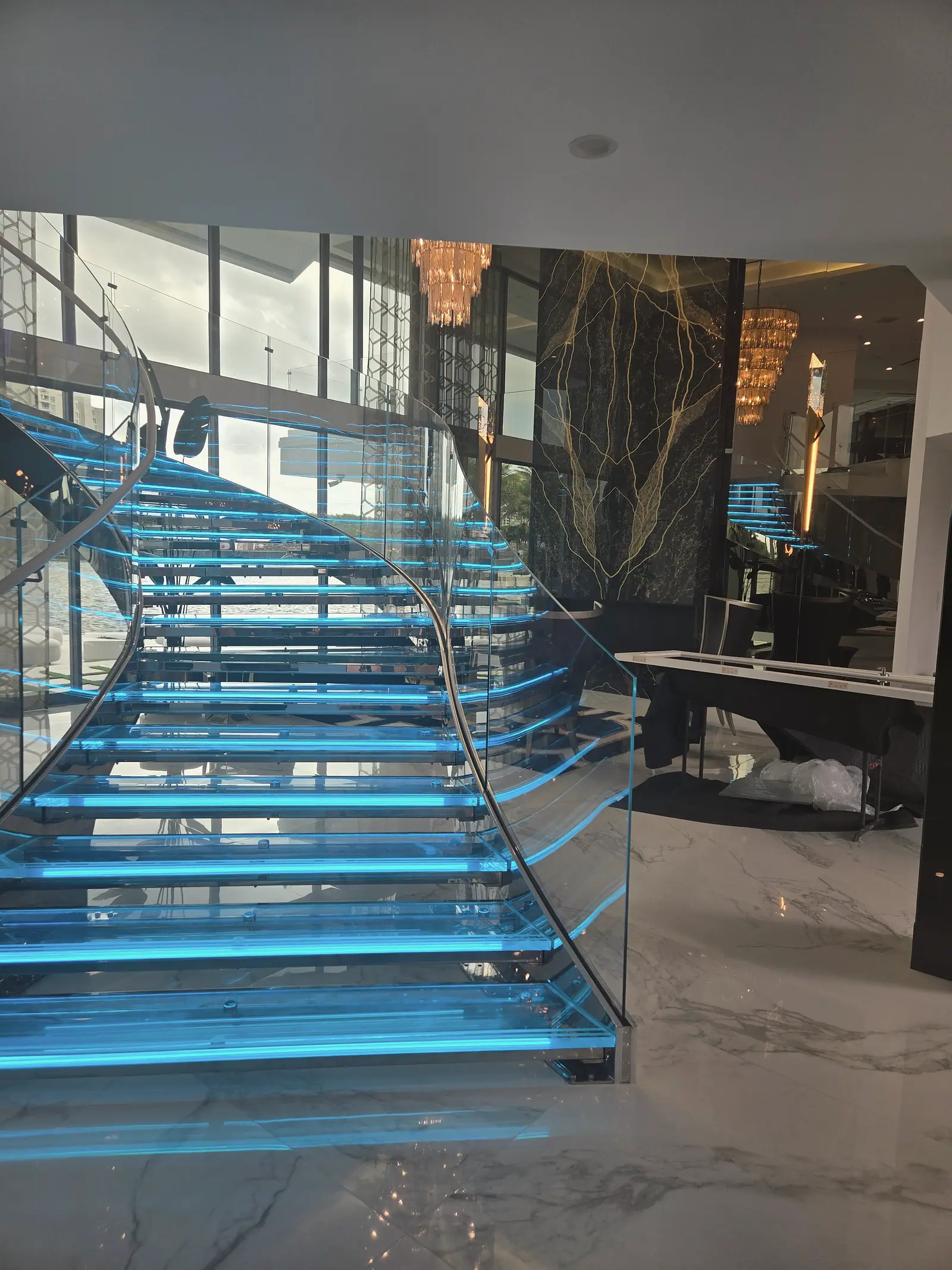 Curved floating glass staircase with blue lit treads and glass railings before a black and gold stone wall