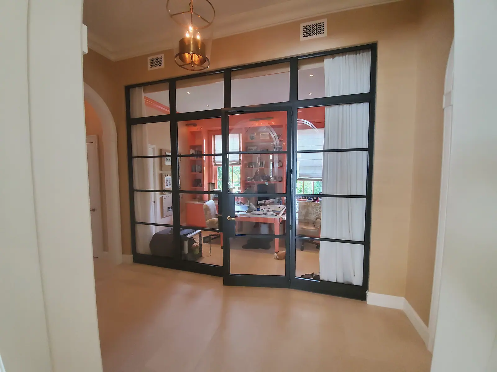 Black steel and glass partition wall with transoms and door into a coral painted office with built in shelves, brass lantern above