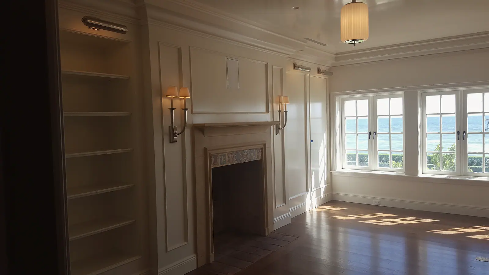 Empty paneled room with a fireplace, built-in shelving, wood floor and windows overlooking the ocean