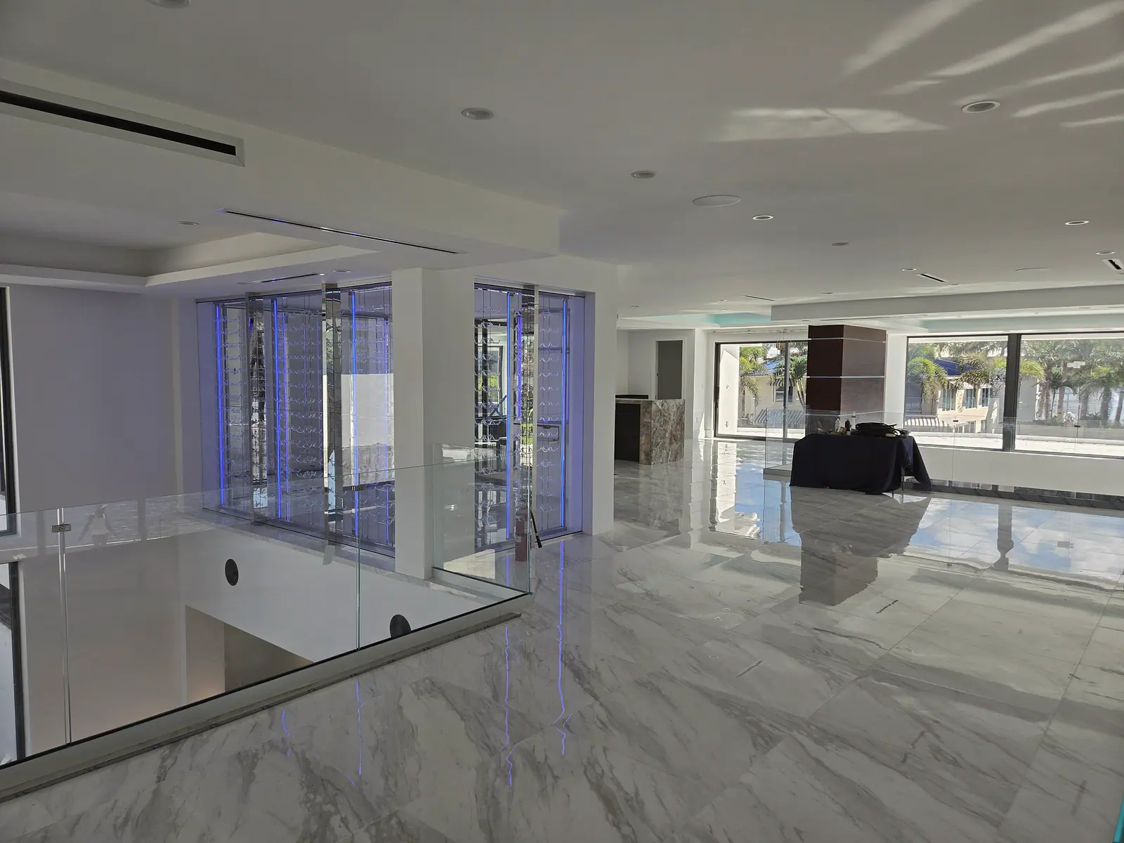 Upper floor with polished white marble tile, frameless glass railing and a glass enclosed wine wall with blue lighting
