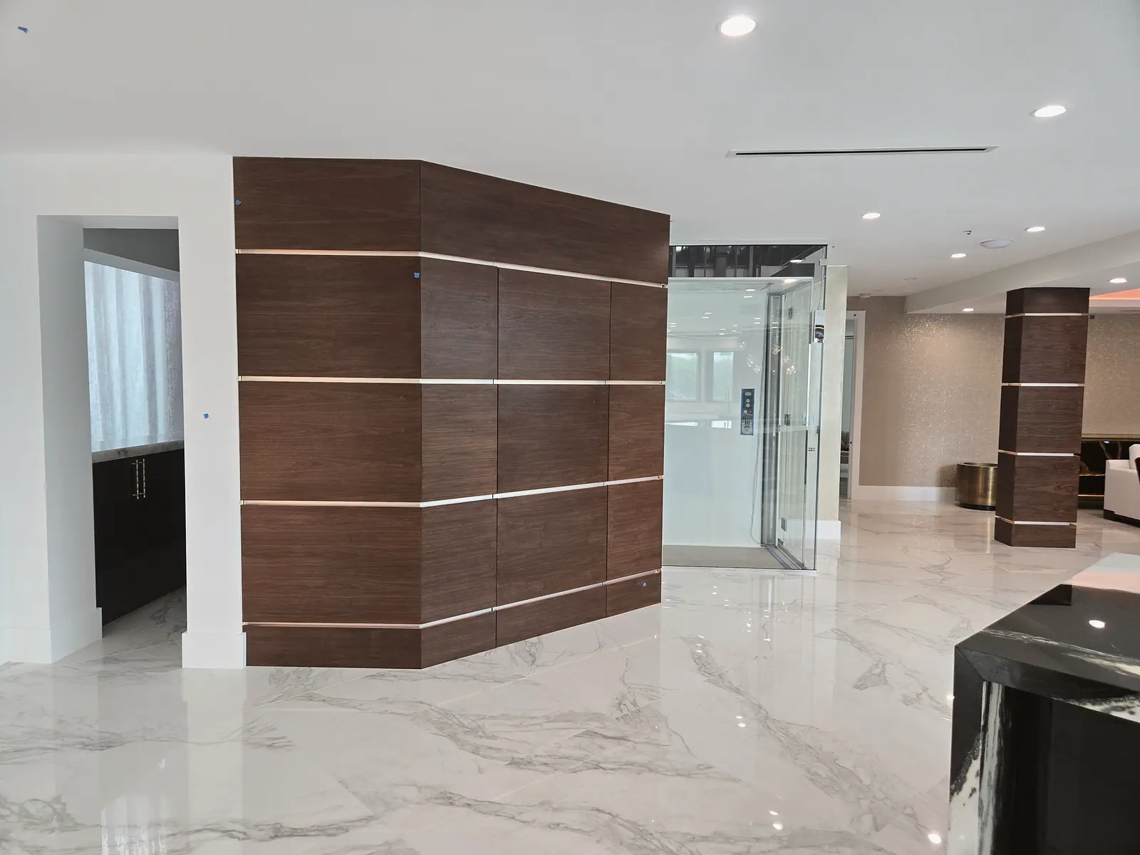 Walnut wall panels with metal reveal strips beside a glass home elevator on white marble floors