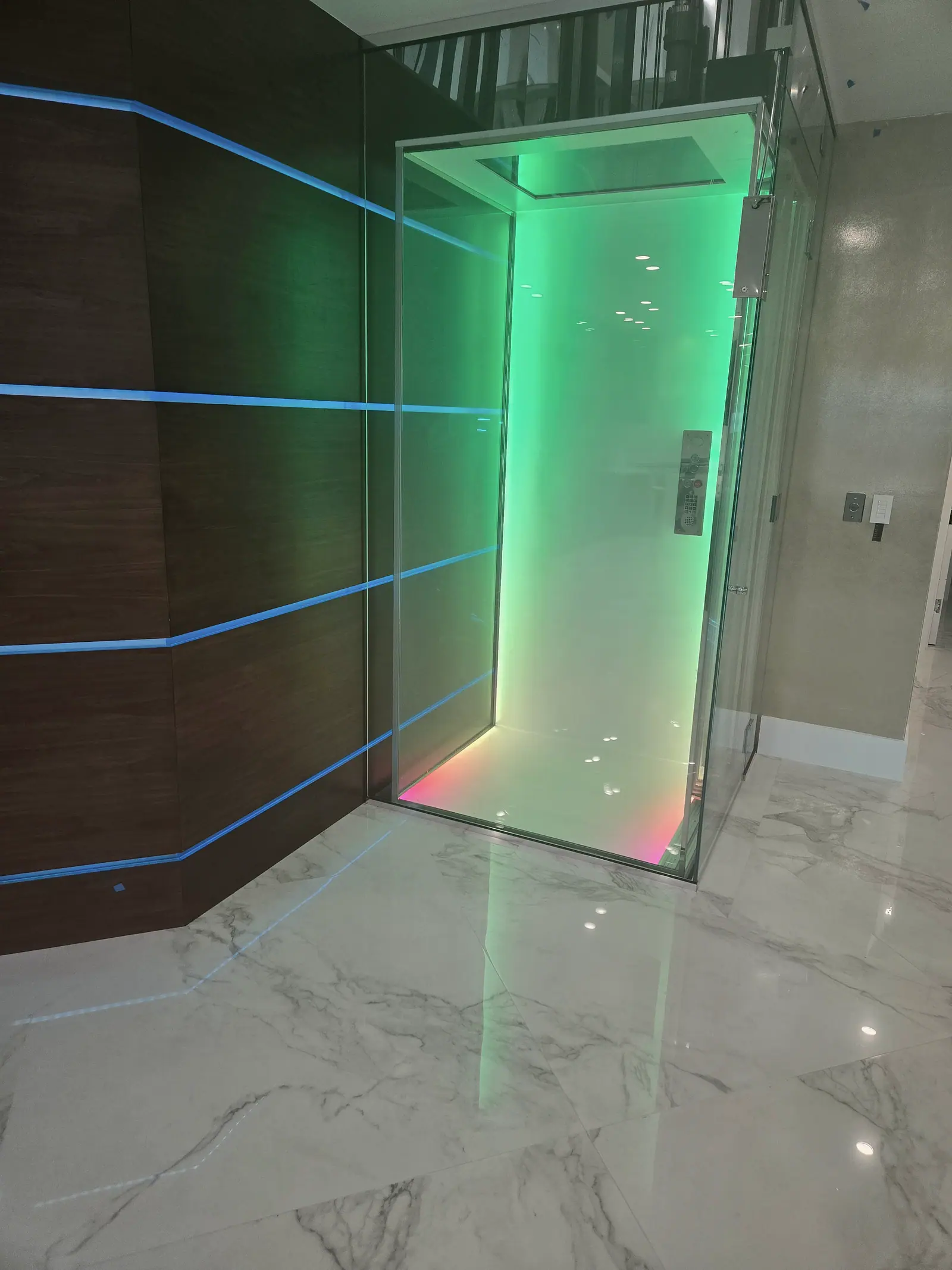 Glass home elevator lit green beside walnut wall panels with blue LED reveal lines