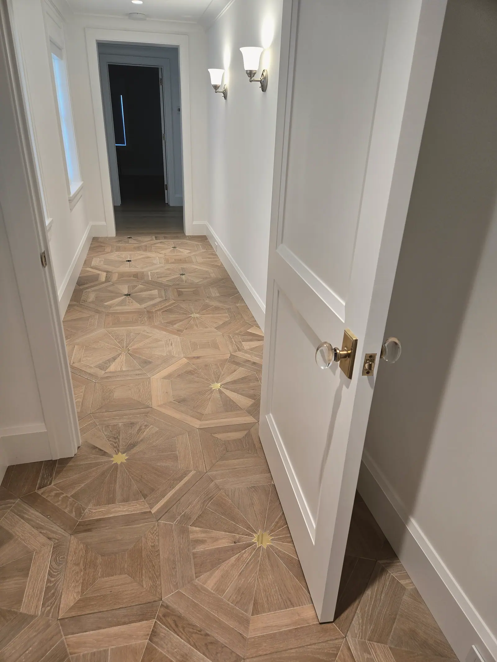 White hallway with wood parquet floor and brass star inlays, wall sconces and a paneled door with glass knobs