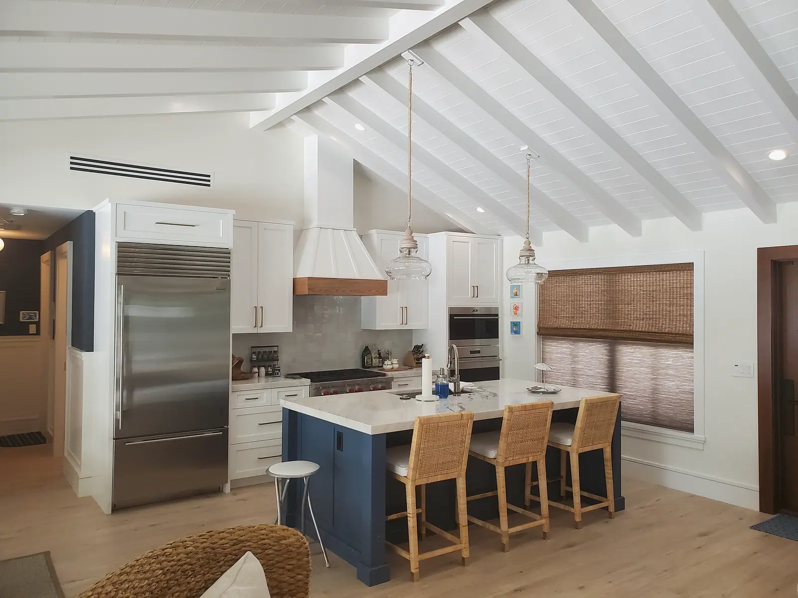 Kitchen with a vaulted white beamed ceiling, white shaker cabinets, a navy island with marble top and rattan stools