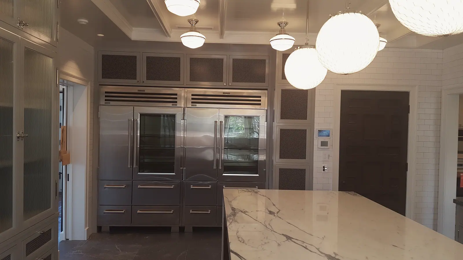 Twin stainless glass-door refrigerators set in gray cabinetry with mesh panels and a marble island
