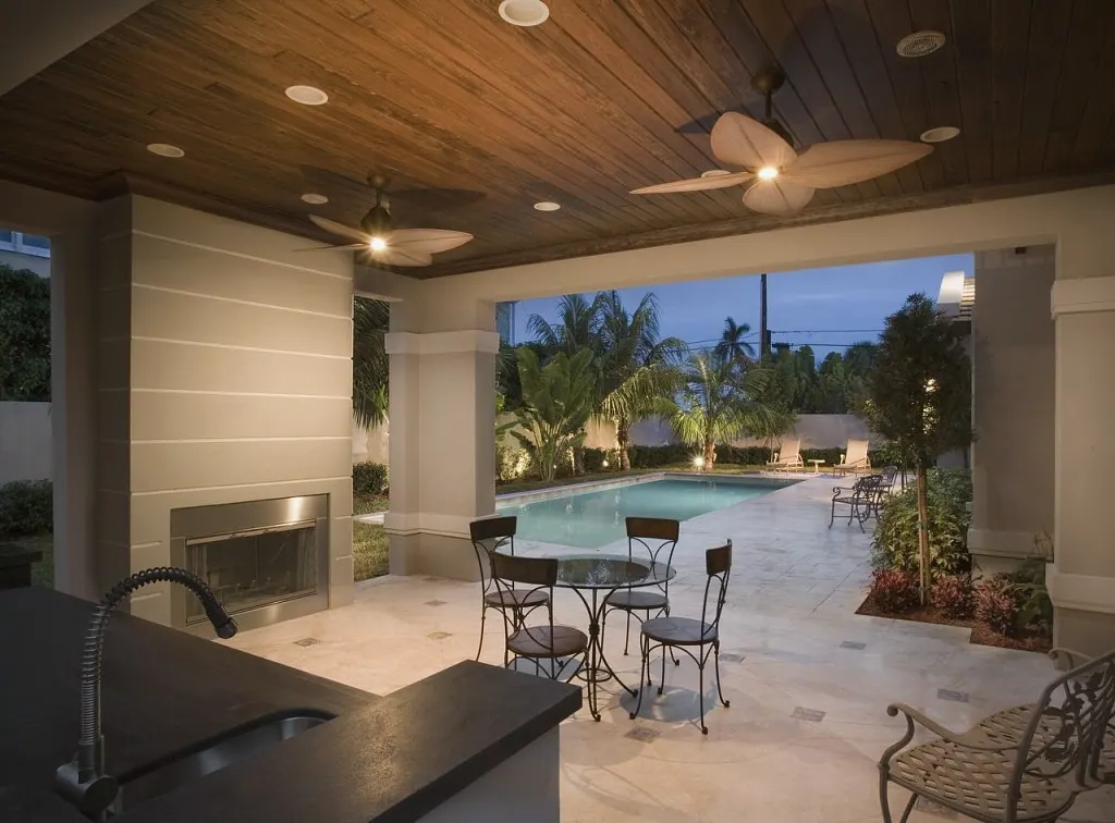 Outdoor kitchen under a wood ceiling with a fireplace, looking out to a lit pool