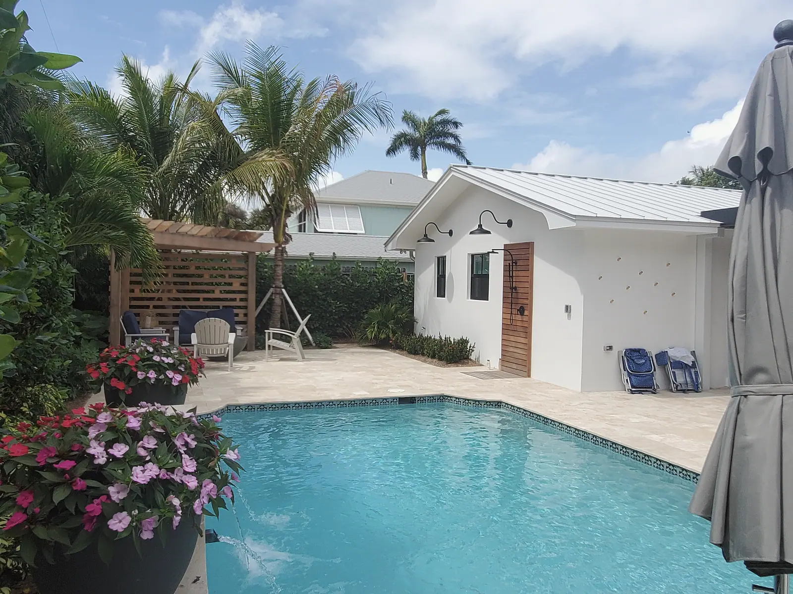 Pool with travertine deck beside a white cabana with standing seam roof and wood slat outdoor shower, cedar pergola seating behind