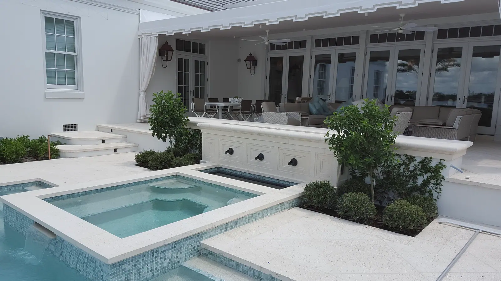 Glass mosaic spa spilling into a pool beside a carved stone fountain wall, with a furnished loggia and French doors behind