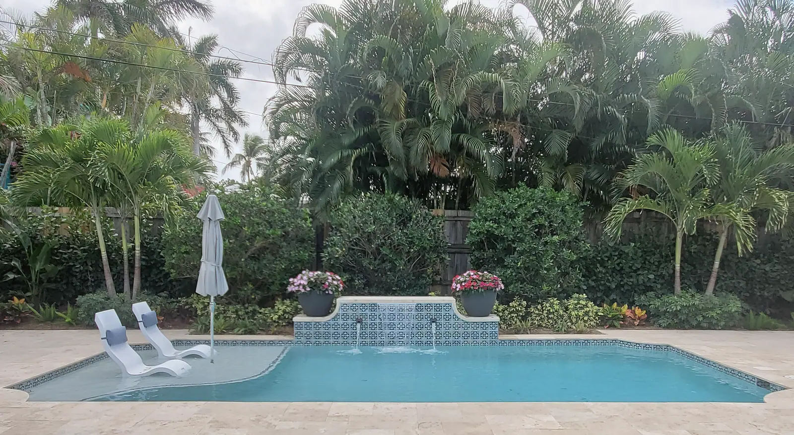 Pool with tanning ledge loungers and a patterned tile fountain wall with spouts and flower urns, framed by palms and a travertine deck