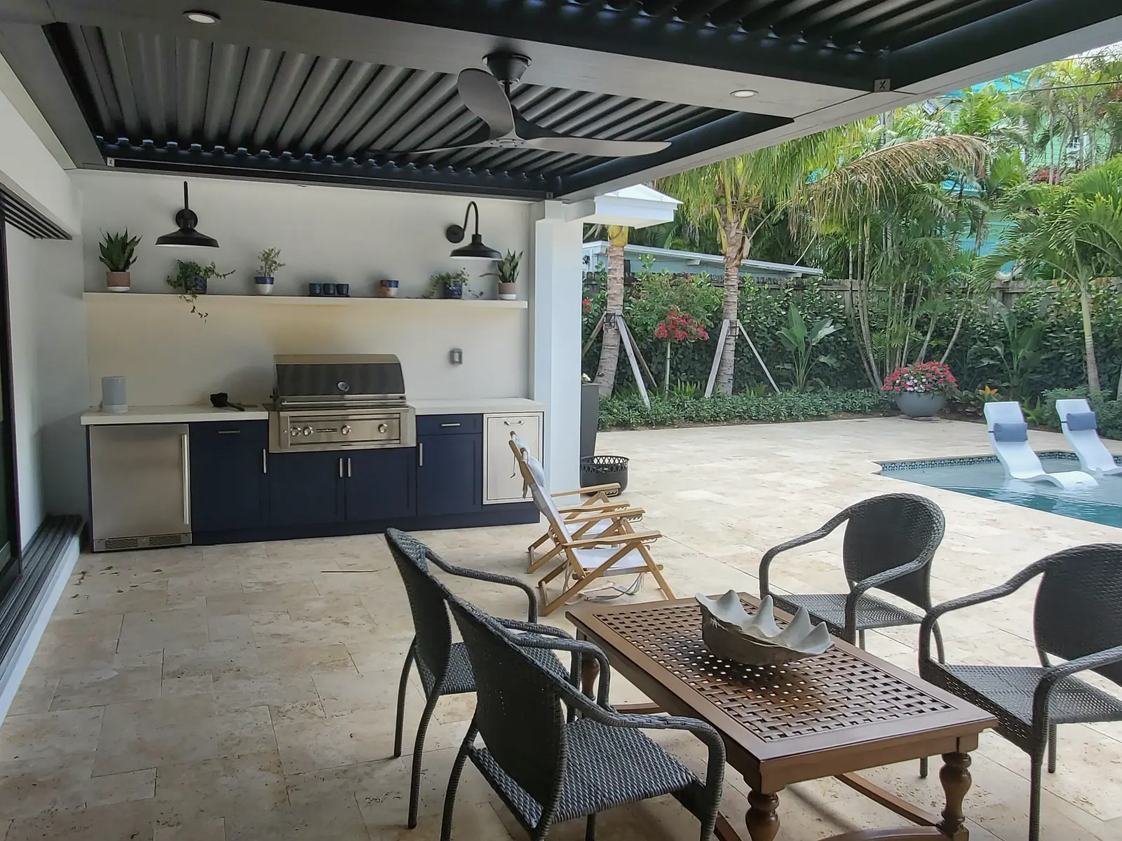 Outdoor kitchen with built-in grill and navy cabinets under a louvered pergola beside a travertine pool deck
