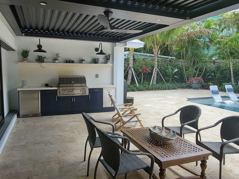 Outdoor kitchen with built-in grill and navy cabinets under a louvered pergola beside a travertine pool deck