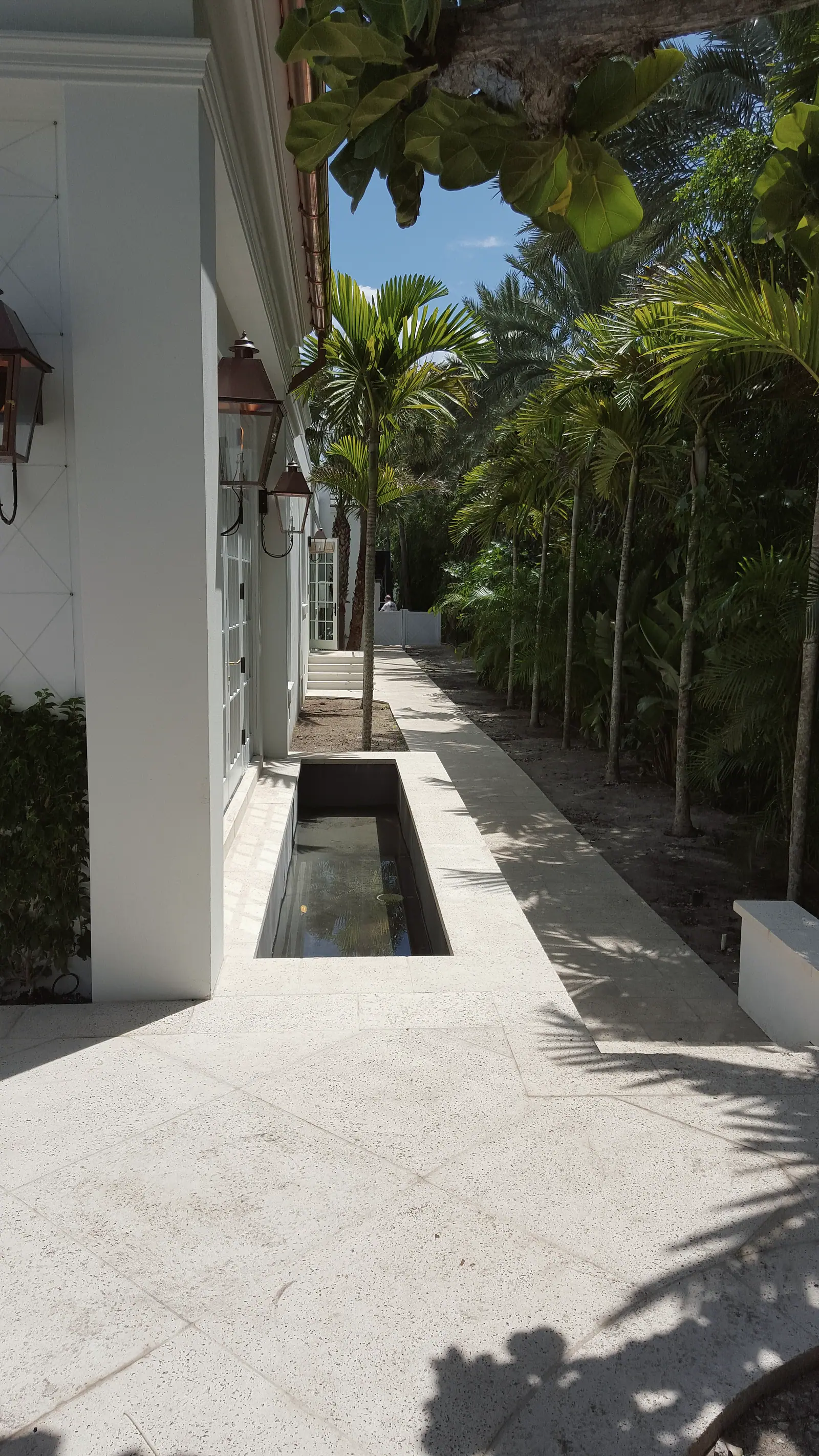 Narrow reflecting pool and keystone walk beside a white house with copper lanterns and a row of palms