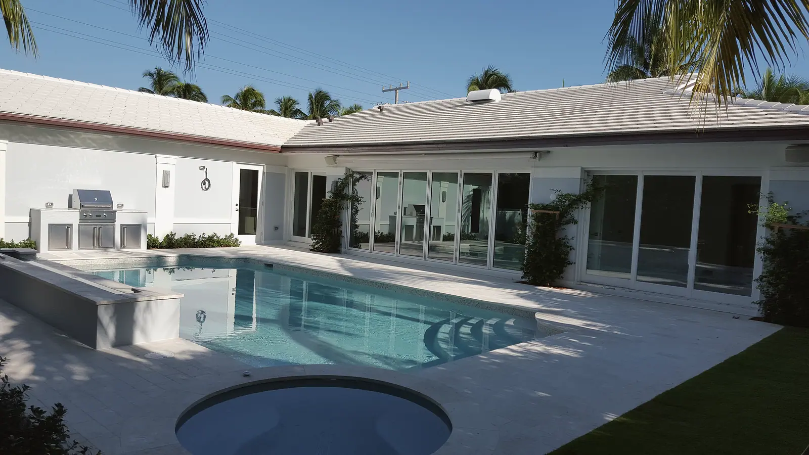 Single story white house with flat tile roof wrapping a pool, round spa and stainless outdoor grill station, with walls of glass doors