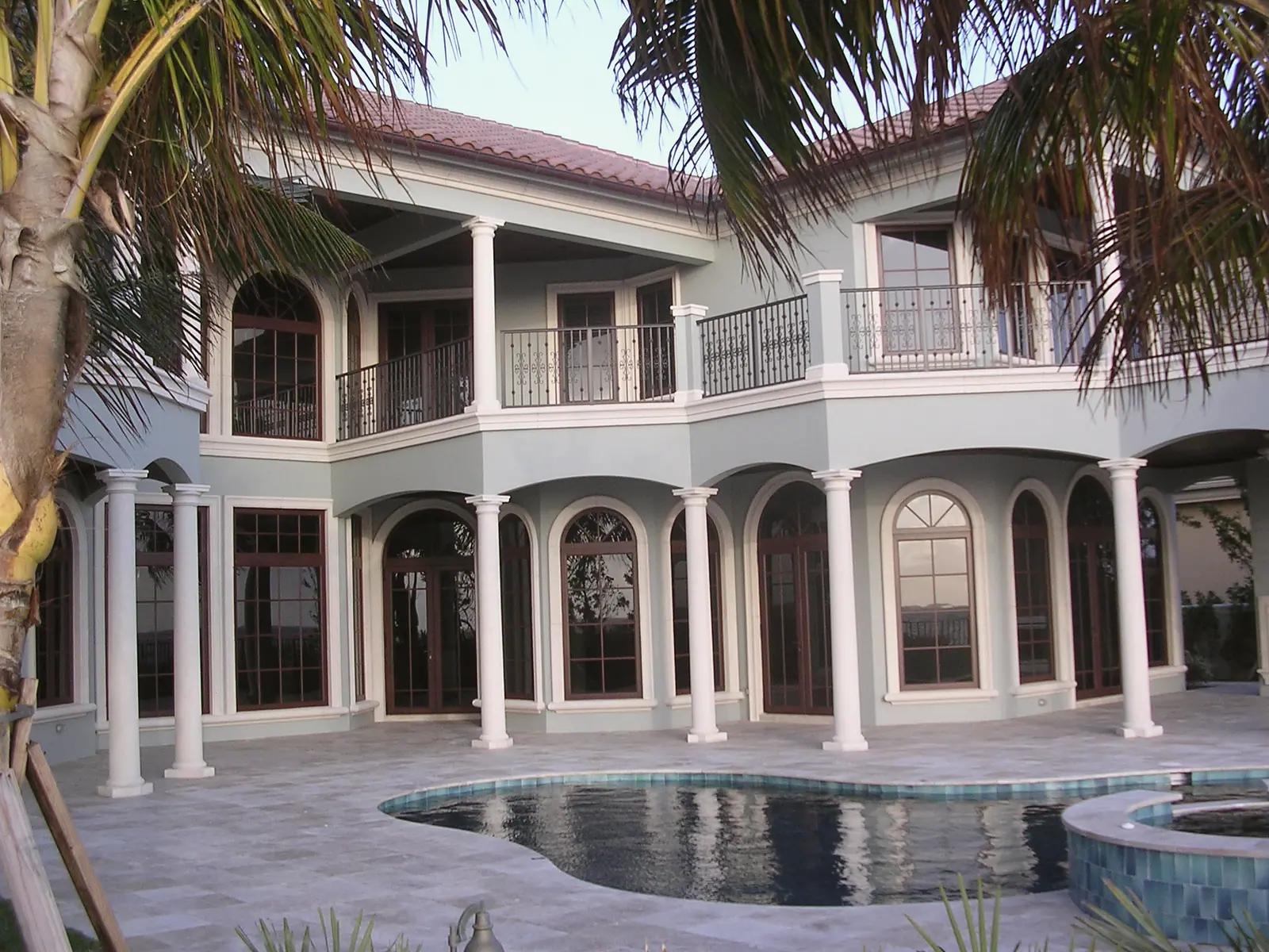 Two-story columned loggia with iron balcony railings above a travertine pool deck with pool and spa