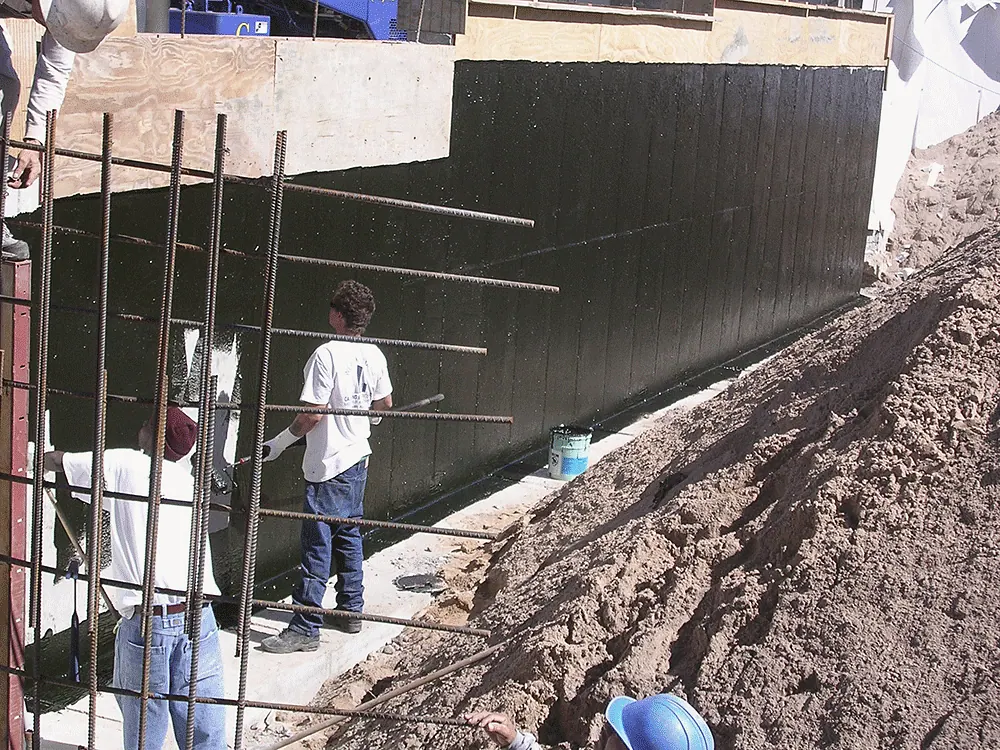 Crew waterproofing a foundation wall, the rebar standing
